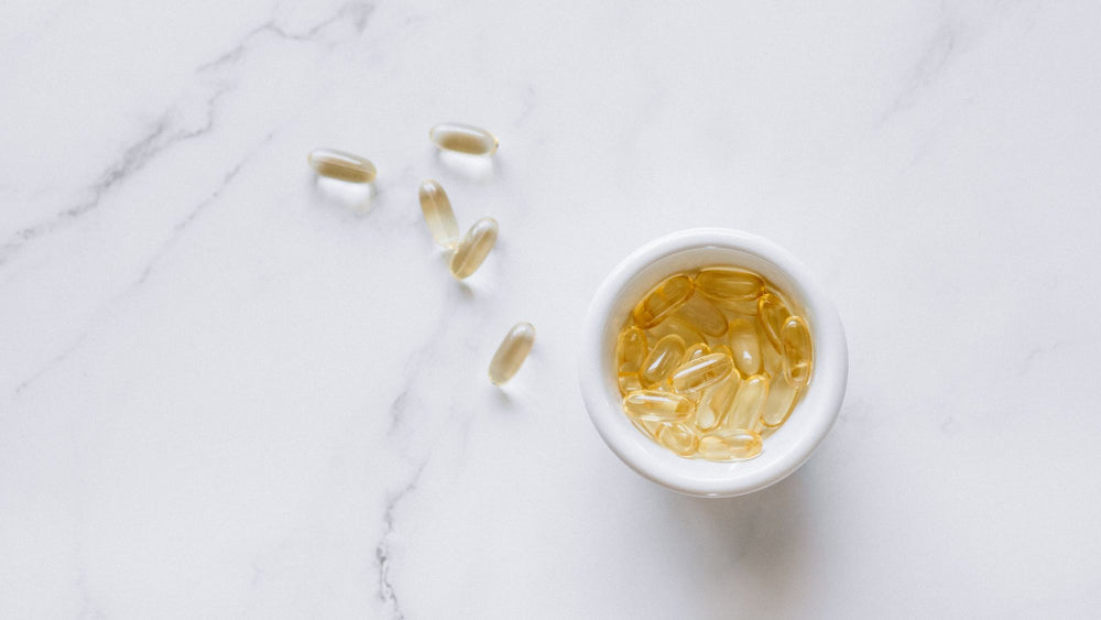 Amber omega-3 softgels in a white ceramic bowl on pale stone, the EPA and DHA carriers photographed as a specimen